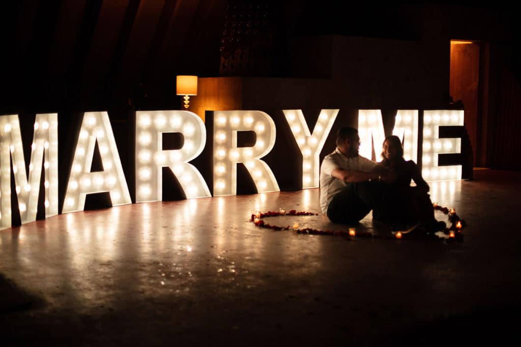 couple sitting in a room with a glowing sign that says "marry me" behind them